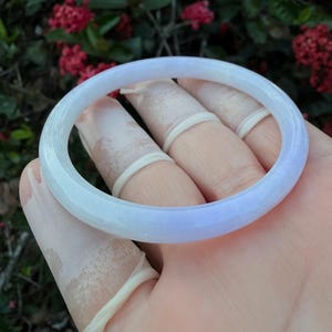Könnte beinhalten: Ein hellblaues und weißes Jade-Armreif-Armband. Die polierte Oberfläche des Armbands reflektiert das Licht und zeigt einen Farbverlauf. Das Armband wird in einer Hand gehalten, mit einem verschwommenen Hintergrund aus roten Blumen und grünem Laub.