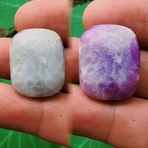 Puede incluir: Se muestran dos piedras preciosas pulidas, una azul claro y otra morada. Las piedras tienen forma rectangular redondeada y un aspecto texturizado y veteado. Las piedras preciosas se sostienen en una mano.