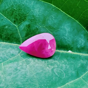 Pode incluir: Uma gema de rubi facetada em forma de pêra, exibindo um tom rosa vibrante. A gema está colocada sobre um fundo de uma grande folha verde texturizada, realçando sua cor e forma. O corte do rubi revela múltiplas facetas.