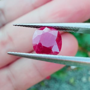 May include: A faceted, cushion-cut ruby gemstone held by tweezers. The ruby is a deep, saturated red color, with multiple facets that reflect light. The tweezers are silver-colored, and the background is blurred green.