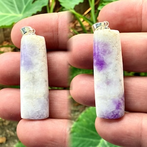 Puede incluir: Un colgante de piedra preciosa blanca y morada con una argolla de plata. La piedra preciosa tiene una textura rugosa y natural y una forma irregular.