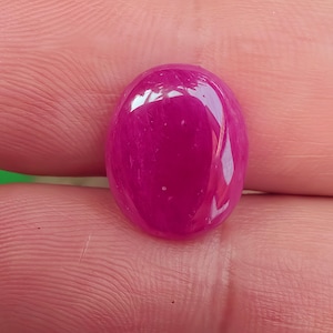 May include: A polished, oval-shaped gemstone in a vibrant magenta hue. The gem has a smooth, glossy surface, reflecting light. The stone is held between fingers, showcasing its size and color.