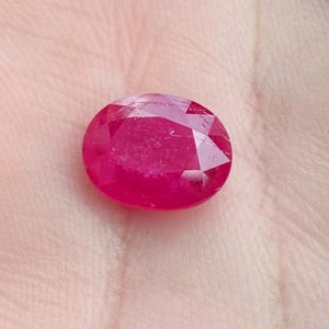 May include: A faceted, oval ruby gemstone. The ruby is a deep red colour with a polished surface. The gemstone is held in a hand, showcasing its size and brilliance. The gem is a symbol of luxury and beauty.