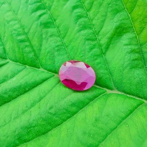 以下が含まれることがあります： 鮮やかなピンクレッドの色合いの、ファセット加工された楕円形のルビーの宝石が、質感のある明るい緑色の葉の上に置かれています。宝石の研磨された表面は光を反射し、きらめく効果を生み出しています。葉の葉脈は、宝石の輝きに自然なコントラストを与えています。