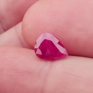 May include: A faceted, pear-shaped ruby gemstone. The gem is a deep, vibrant red with lighter pink and white facets. The ruby is held in a hand, showcasing its size and color.
