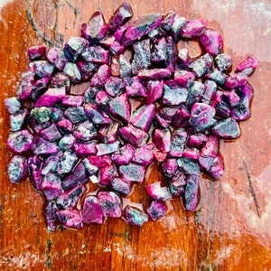 May include: A collection of small, rough, purple and green gemstones, scattered on a wooden surface.