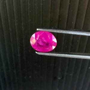 May include: A faceted, oval-shaped ruby gemstone, held by a pair of tweezers. The ruby is a vibrant, deep pink color, with visible internal inclusions. The background is a dark, textured surface.