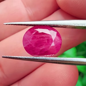 以下が含まれることがあります： ピンセットの金属製の先端で挟まれた、オーバルカットの鮮やかなピンクがかった赤色の宝石。石の表面はファセットカットが施されており、内部のインクルージョンが確認でき、全体的に深く鮮やかな色合いで磨き上げられた仕上がりとなっています。
