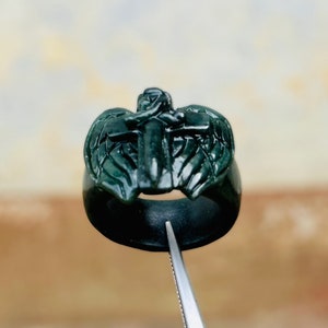 May include: A dark green ring with a detailed carving of a cross with wings. The ring is made of a smooth, polished material.