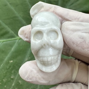 May include: A white, carved skull made of polished stone. The skull has a detailed face with eye sockets, a nose, and a wide smile.