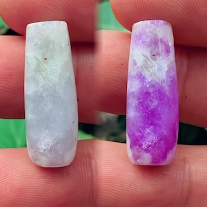 Puede incluir: Dos piedras preciosas rectangulares y pulidas. Una es de color lavanda pálido, la otra es de un púrpura vibrante con vetas blancas. Ambas se sostienen entre los dedos, mostrando sus superficies lisas y translúcidas. Las piedras tienen una forma ligeramente cónica.