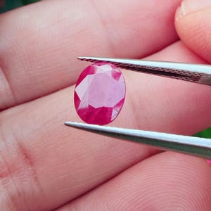 May include: A faceted, oval-shaped ruby gemstone held by tweezers. The ruby displays a deep red color with multiple facets reflecting light. The gemstone is the focal point, showcasing its clarity and brilliance.