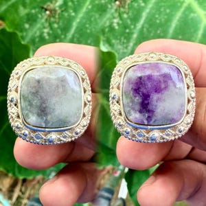 Könnte beinhalten: Zwei Silberringe mit quadratischen, facettierten Edelsteinen. Der Ring links hat einen hellgrauen Stein, während der Ring rechts einen lila und grauen Stein hat.