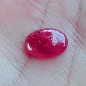 May include: A polished, oval-shaped, ruby-red gemstone. The gem has a smooth, glossy surface with light reflections. The stone is set against a light-colored background.