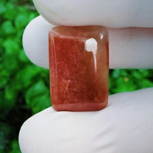 May include: A rectangular, polished gemstone with a smooth, cabochon-like surface. The stone displays a deep, translucent reddish-orange hue with internal cloudy inclusions and a subtle, reflective sheen across its flat, rounded edges.
