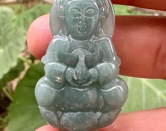 May include: A jade green pendant carved in the shape of a seated Buddha figure. The pendant is detailed with a serene facial expression and intricate folds in the clothing.