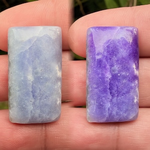 Puede incluir: Dos piedras preciosas rectangulares, una azul claro y otra púrpura, se muestran una al lado de la otra. Las piedras tienen una superficie pulida y lisa con patrones de vetas naturales. Las piedras preciosas se sostienen entre los dedos.