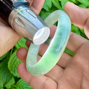 Könnte beinhalten: Ein hellgrünes Jade-Armreif-Armband. Das durchscheinende Armband wird mit einer Taschenlampe untersucht. Der Jade hat eine glatte, polierte Oberfläche und verschiedene Grüntöne. Der Hintergrund ist verschwommen, mit grünem Laub.