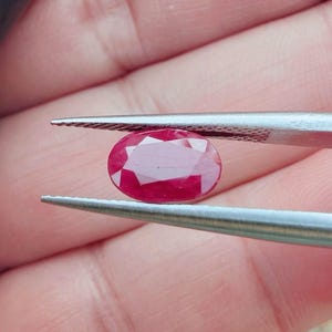 Peut inclure: Un rubis facetté de forme ovale, maintenu par une pince. Le rubis affiche une couleur rouge profond avec de multiples facettes réfléchissant la lumière. La pierre précieuse est un point central, mettant en valeur sa clarté et sa taille.