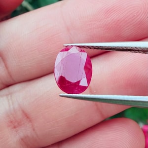 May include: An oval-cut, faceted ruby gemstone. The ruby is a deep, vibrant red color and is held by tweezers. The gemstone's facets reflect light, creating a sparkling effect. The background is blurred, with hints of green and pink.