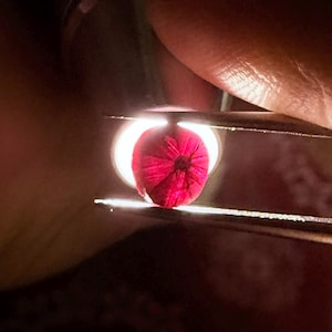 May include: A close-up of a vivid red gemstone held in tweezers. The gemstone is lit from behind, highlighting its complex internal structure and deep colour. The tweezers are silver, and the background is dark.