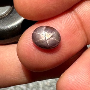 May include: A close-up of a dark purple gemstone with a star-shaped pattern. The gemstone is round and has a smooth, polished surface.