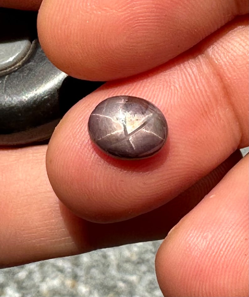 May include: A close-up of a dark purple gemstone with a star-shaped pattern. The gemstone is round and has a smooth, polished surface.