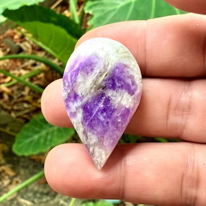 Puede incluir: Un cabujón de amatista en forma de lágrima, de color púrpura y blanco, con un acabado natural y en bruto.