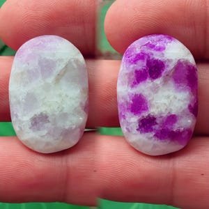 Puede incluir: Dos piedras preciosas ovaladas, una lavanda pálida y blanca, la otra morada vibrante y blanca. Las piedras tienen una superficie pulida y lisa con apariencia veteada. Las piedras preciosas se sostienen entre los dedos.