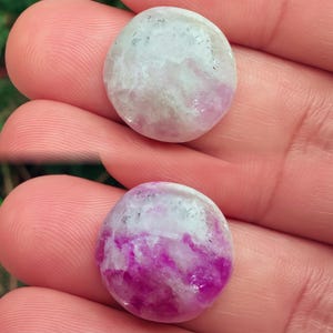 Puede incluir: Dos piedras preciosas redondas y pulidas con una mezcla de tonos blancos y rosas. La piedra superior tiene un rosa más claro, mientras que la piedra inferior es de un magenta más profundo. Las piedras se sostienen en una mano, mostrando su forma suave y redondeada.
