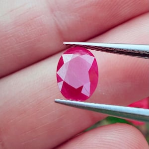 May include: A faceted, oval-shaped ruby gemstone held by tweezers. The ruby is a deep, vibrant red color, with multiple facets catching the light. The gemstone is held against a blurred background.