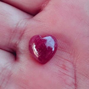 Op de afbeelding: Een kleine, hartvormige, gepolijste edelsteen in een dieprode kleur. De steen heeft een glad, glanzend oppervlak en wordt in een hand gehouden. De hartvorm is duidelijk gedefinieerd, met afgeronde randen en een lichte inkeping aan de bovenkant.