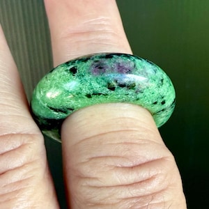 May include: A polished, oval-shaped ring made of green and black stone, with flecks of red and purple. The ring is worn on a finger, showcasing its smooth, rounded design. The gemstone ring is a unique piece of jewelry.