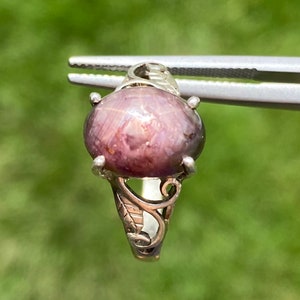 Op de afbeelding: Een zilveren ring met een grote ovale, paarse ster-saffier edelsteen. De ring heeft een delicaat, versierd ontwerp.