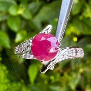 Op de afbeelding: Een zilveren ring met een grote, ronde, rode robijn in het midden. De ring heeft een gedraaide band met kleinere diamanten aan de zijkanten.