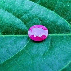 以下が含まれることがあります： 鮮やかな緑色の葉の上に、楕円形にカットされたルビーの宝石が置かれています。ルビーは、内部のファセットが明るく、深いピンク色をしており、光の遊びを作り出しています。葉の質感と葉脈が見えます。