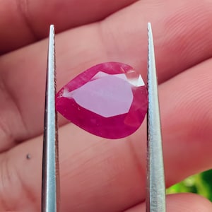 May include: A pear-shaped, faceted ruby gemstone held by tweezers. The ruby is a deep red colour with a lighter pink interior. The tweezers are silver-coloured and the background is blurred.