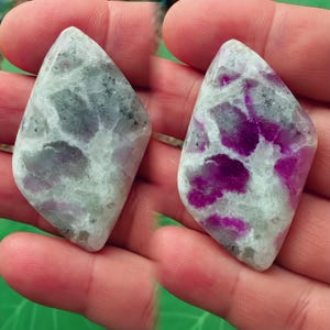 Puede incluir: Dos piedras preciosas pulidas con forma de diamante. Presentan una mezcla de colores blanco, gris y magenta. Las piedras se sostienen en una mano, mostrando sus patrones y texturas únicos.