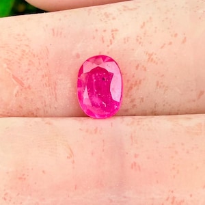 May include: A faceted, oval-cut ruby gemstone. The gem is a vibrant, saturated pink-red color, with visible internal inclusions. The shape is elongated, with a polished surface reflecting light.