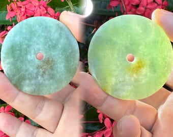 Könnte beinhalten: Zwei Jade-Anhänger, einer hellgrün und einer limettengrün, jeweils mit einem zentralen Loch. Die Anhänger sind rund und glatt, mit natürlichen Farb- und Texturvariationen. Der Hintergrund zeigt rote Blumen.