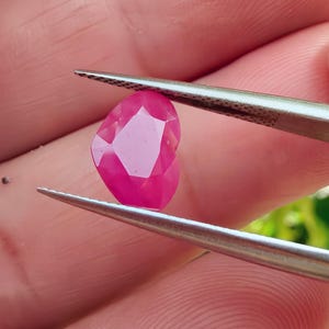 以下が含まれることがあります： ピンセットで保持された、ファセット加工されたハート型のルビー。ルビーは鮮やかなピンク色で、複数のファセットが光を捉えています。ピンセットは銀色で、背景はぼやけています。