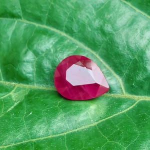 May include: A faceted, pear-shaped ruby gemstone, displaying a deep red hue, rests on a vibrant green leaf. The gemstone's cut reveals multiple facets, reflecting light. The leaf's texture and veins are visible, creating a natural contrast.