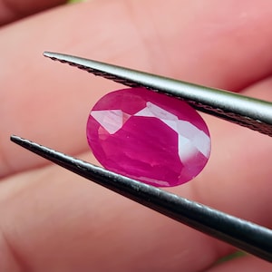 May include: A faceted, oval-shaped ruby gemstone held by tweezers. The ruby is a vibrant pinkish-red color, with multiple facets reflecting light. The tweezers are silver-colored and hold the gemstone against a blurred background.