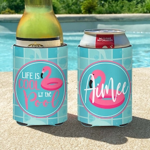 May include: Two blue and white can coolers with a pink flamingo and inflatable ring design. One cooler says "Life is Cool by the Pool" and the other says "Aimee".