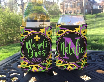 May include: Two Mardi Gras themed can coolers. One cooler has the text "Mardi Gras 2025" and the other has the letter "N" and the name "Natalie". Both coolers have a black background with purple, green, and gold beads and masks.