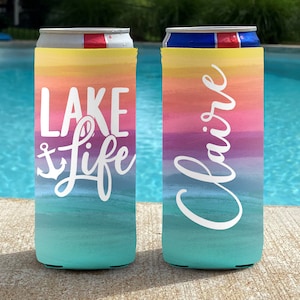 May include: Two colorful can coolers with a watercolor design. One cooler has the words "Lake Life" and an anchor graphic. The other cooler has the name "Claire" in white script. The coolers have a gradient of yellow, orange, pink, purple, and teal.