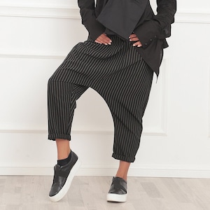 May include: A black ensemble featuring a long-sleeved shirt with a unique asymmetrical design and a matching crossbody bag. The outfit is paired with black and white striped harem pants and black sneakers. The overall style is modern and chic.