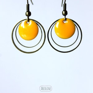 Può includere: Un paio di orecchini gialli e color ottone. Ogni orecchino presenta un pendente circolare giallo brillante, sospeso all'interno di due cerchi concentrici color ottone. Gli orecchini pendono da un gancio e una molla color ottone. Gli orecchini sono su sfondo bianco.
