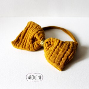 Puede incluir: Diadema con lazo amarillo mostaza de tela texturizada. El lazo está sujeto a una fina banda elástica a juego. Un pequeño adorno dorado está en el centro del lazo. La diadema está sobre una superficie blanca.