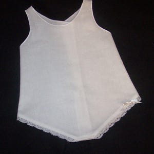 Puede incluir: Una prenda blanca sin mangas con cuello redondo y dobladillo asimétrico acampanado. Delicados bordes de encaje bordean el dobladillo, y hay un pequeño lazo. La tela parece ligera y semitransparente. Es probable que sea un vestido de bebé.